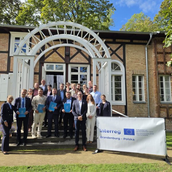 Group of project partners in front of the entrance to the Schweizerhaus Seelow; sunny day, pergola, house, people, banners with sponsorship logo
