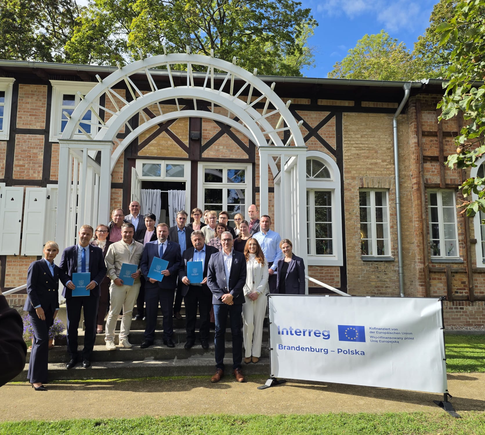 Gruppe von Projektpartnern vor dem Eingang des Schweizerhaus Seelow; sonnig, Pergola, Haus, Menschen, Banner mit Förderlogo